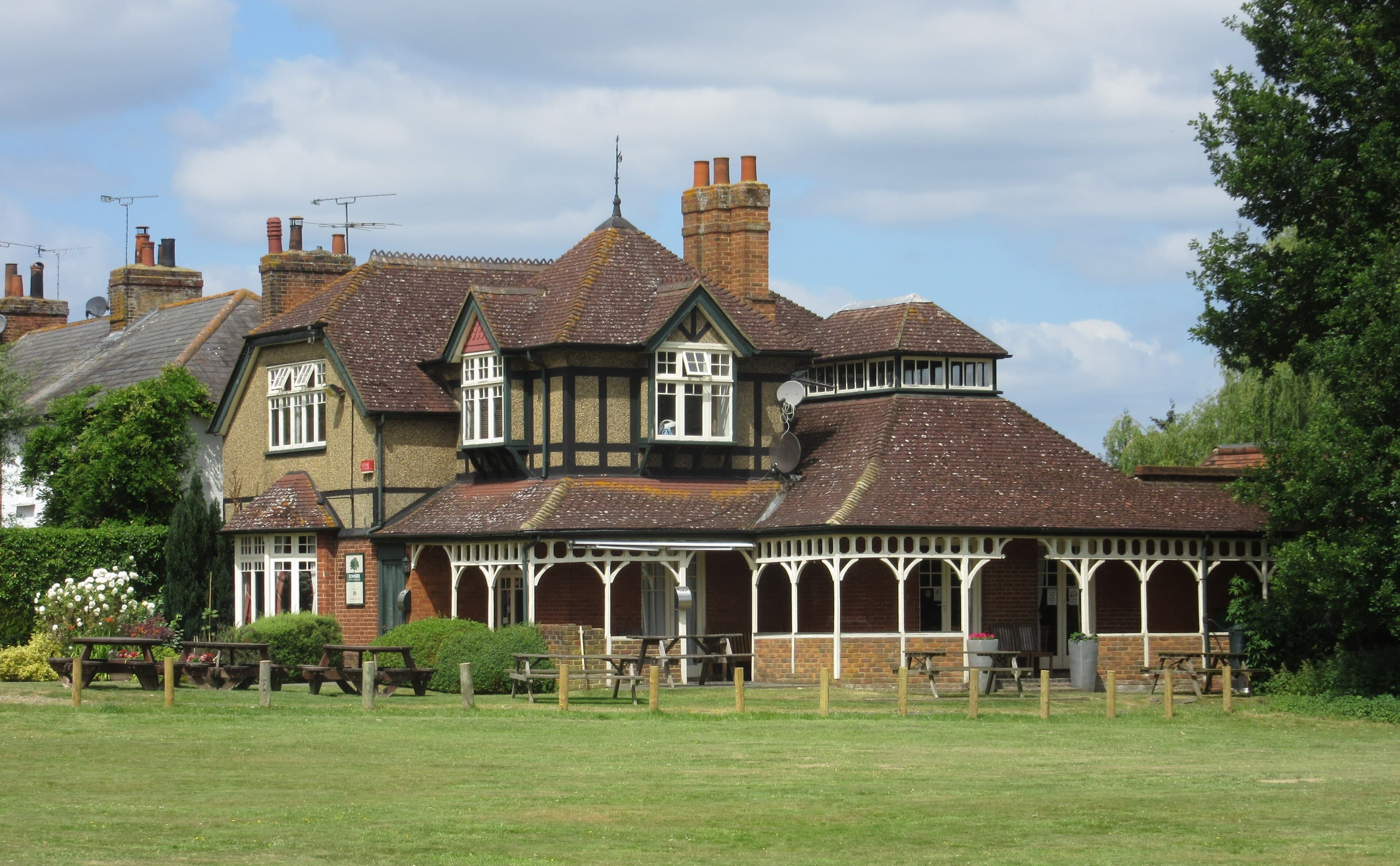 Edwardian brick and tile-hung pub with a white verandah and picnic benches overlooking a village green on a sunny day